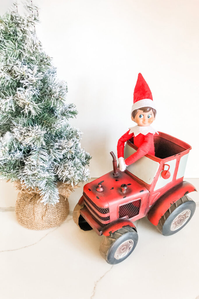 In this image, an elf on the shelf doll is riding in a decorative red tractor next to a mini decorative evergreen tree. This is one of the many elf on the shelf ideas for home included in this post.