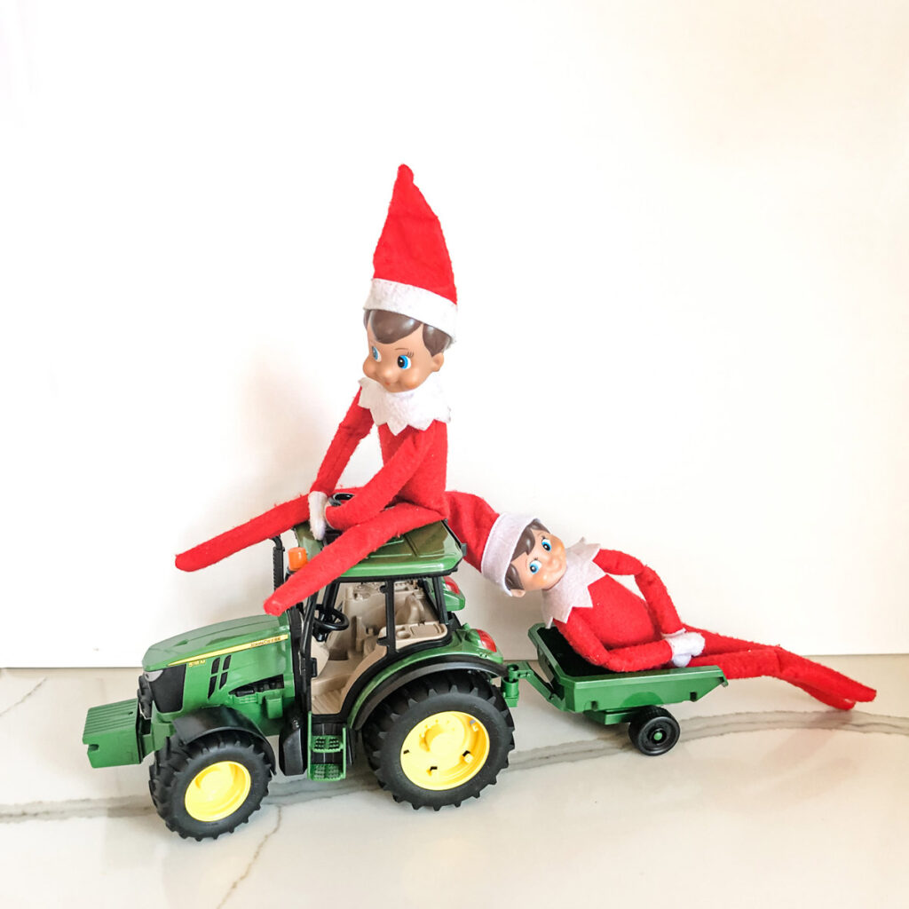 In this image, an elf on the shelf doll is sitting on top of a toy tractor. He is pulling a cart behind him with a second elf on the shelf in the cart. This is one of the many elf on the shelf ideas for home included in this post.