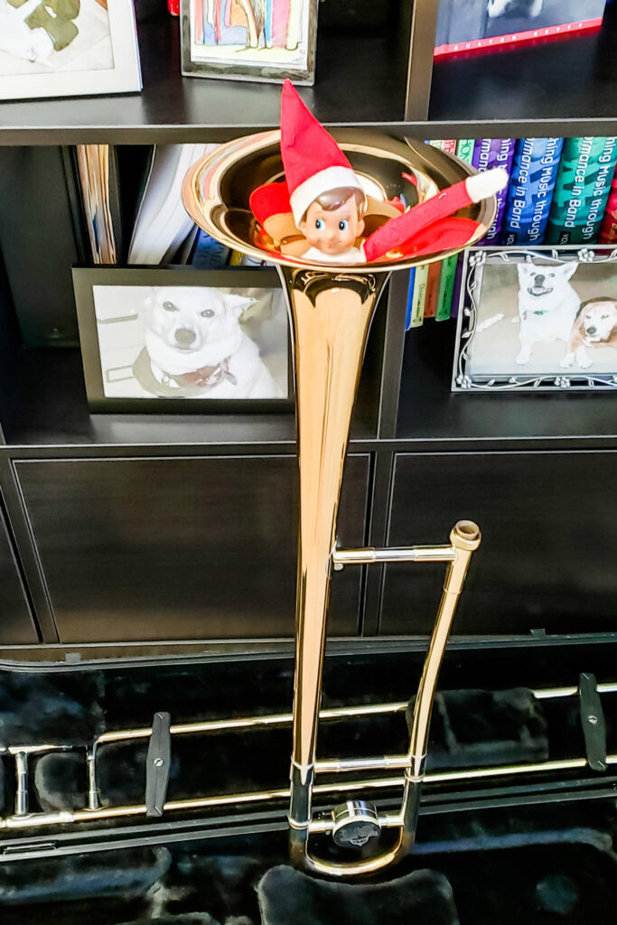 In this image, an elf on the shelf doll is stuck inside of a trombone bell. This is one of the many elf on the shelf ideas for home included in this post.