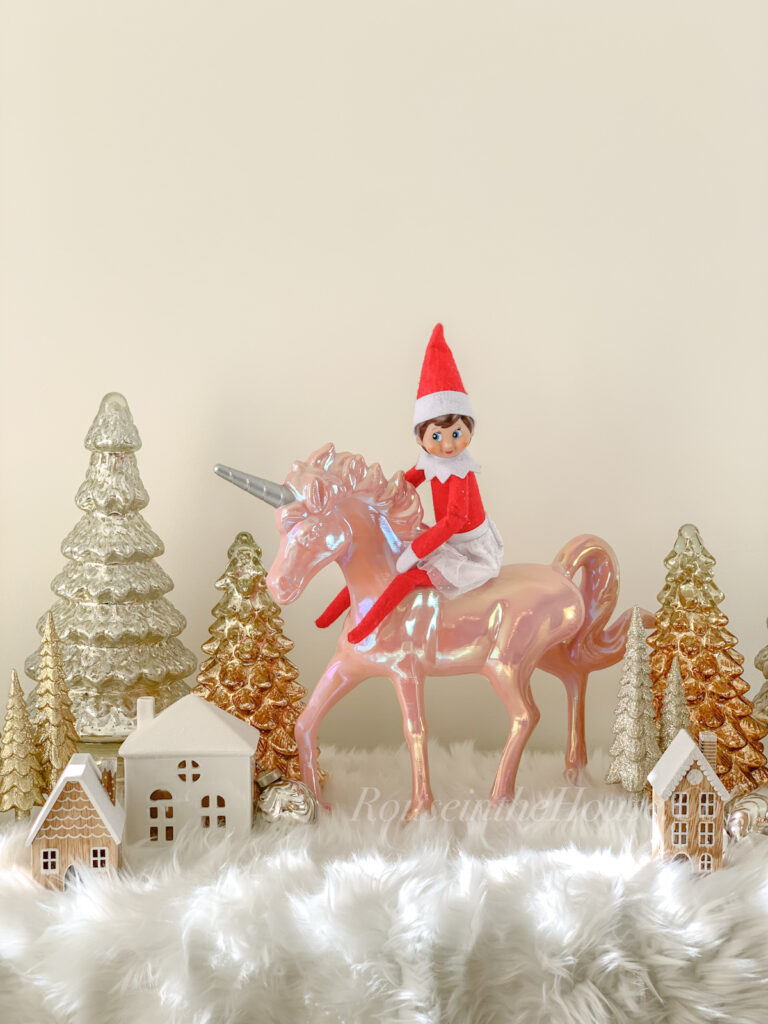 This image shows an Elf on the Shelf doll riding a unicorn toy. She is surrounded by decorative trees and houses. This is one of the many elf on the shelf ideas for home included in this post. Use a unicorn, horse, elephant, etc. toy to create this magical elf on the shelf scene. This is one of the 50+ elf on the shelf ideas for home included in this round up. Plus, you’ll get a set of free Elf on the Shelf planning printables. This includes a blank elf on the shelf calendar, list of elf on the shelf ideas, printable elf on the shelf notes, a fillable elf on the shelf note set, and more.
