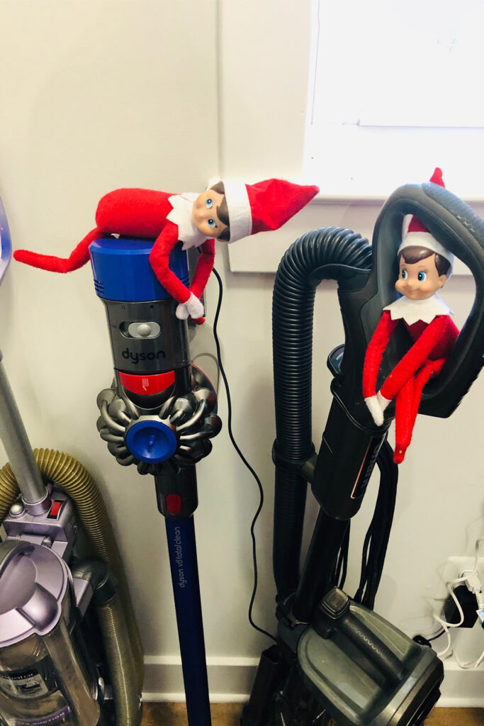 In this picture, there are 2 elf on the shelf dolls sitting on vacuums. This is one of the many elf on the shelf ideas for home included in this post.