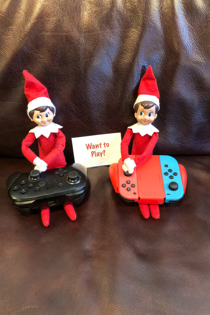 In this image there are 2 elf on the shelf dolls holding Nintendo switch video game controllers. There is a note that says want to play? This is one of the many elf on the shelf ideas for home included in this post.