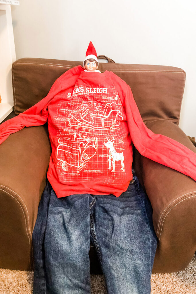 This image shows an elf on the shelf doll with its body stuck inside of a kid’s shirt. It is made to look like he is wearing the shirt and the jeans that are also pictured. This is one of the many elf on the shelf ideas for home included in this post.