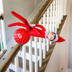 In this image, an elf on the shelf is sitting on a ball that is hanging from a light fixture. This is one of the many elf on the shelf ideas for home included in this post.