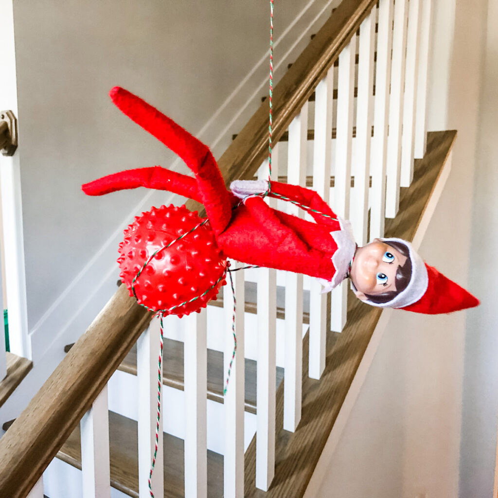 In this image, an elf on the shelf is sitting on a ball that is hanging from a light fixture. This is one of the many elf on the shelf ideas for home included in this post.