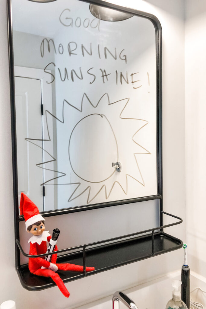 In this image, there is an elf on the shelf sitting on a shelf holding a dry erase marker. Above him is a mirror with good morning sunshine! Written on it with a sun drawn on the mirror, too. This is one of the many elf on the shelf ideas for home included in this post.