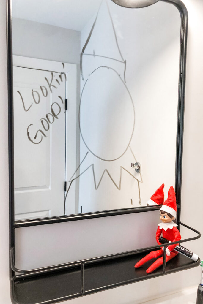 In this image, there is an elf on the shelf sitting on a shelf holding a dry erase marker. Above him is a mirror with lookin good! Written on it with an elf body drawn on the mirror, too. This is one of the many elf on the shelf ideas for home included in this post. This is one of the many elf on the shelf ideas for home included in this post.
