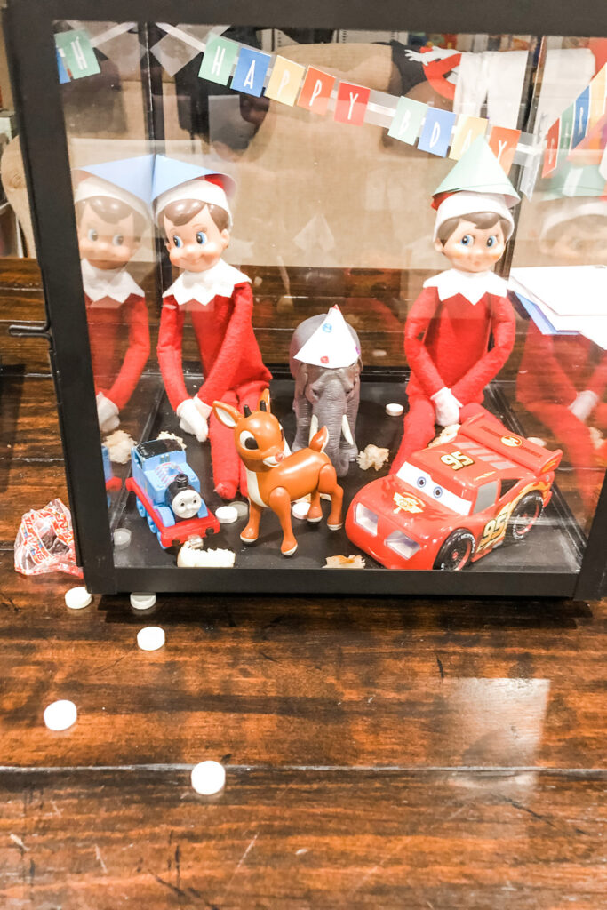 In this image, there is are 2 elf on the shelf dolls with some other small toys pretending to have a birthday party. Behind them is a mini Happy birthday banner, And they are wearing party hats. This is one of the many elf on the shelf ideas for home included in this post.