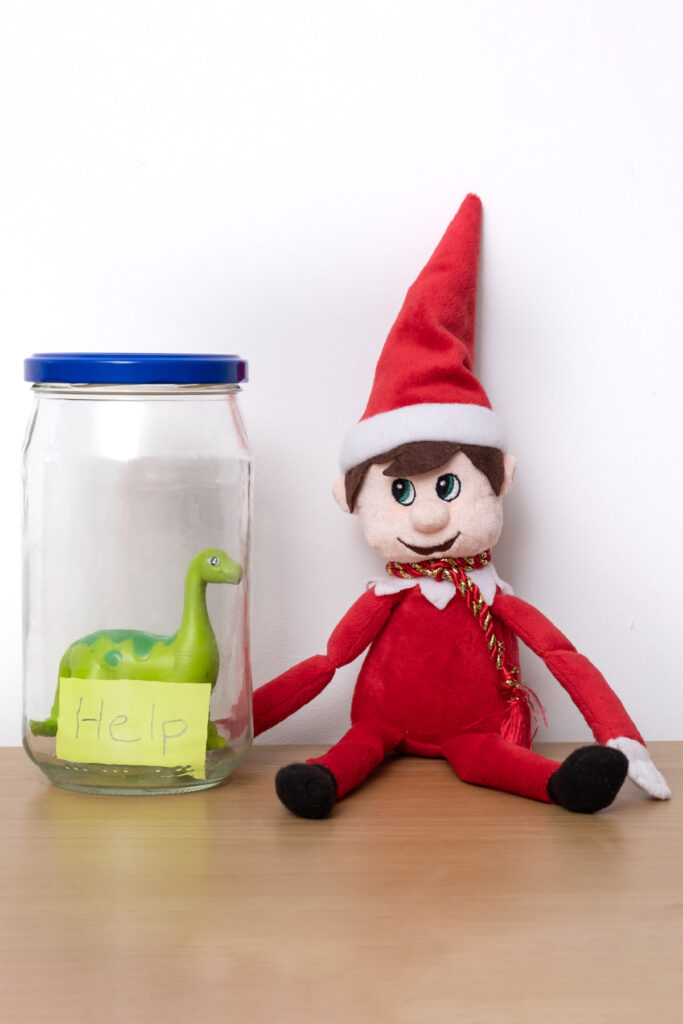 In this image, an elf doll is sitting next to a toy dinosaur trapped inside of a glass jar. The jar has a note on it that says help. This is one of the many elf on the shelf ideas for home included in this post.