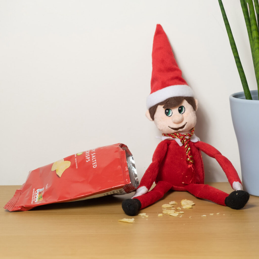 In this image, an elf doll is sitting next to an open bag of chips. There are crumbs all over the table. This is one of the many elf on the shelf ideas for home included in this post.