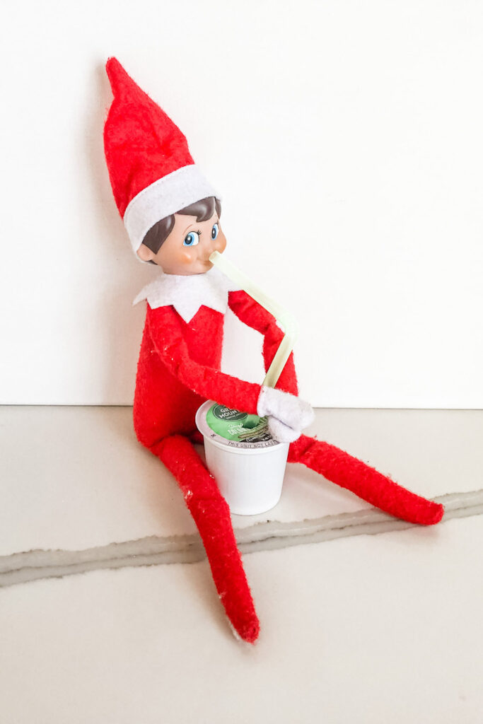 In this image, an elf is pretending to drink out of a k-cup. This is one of the many elf on the shelf ideas for home included in this post.