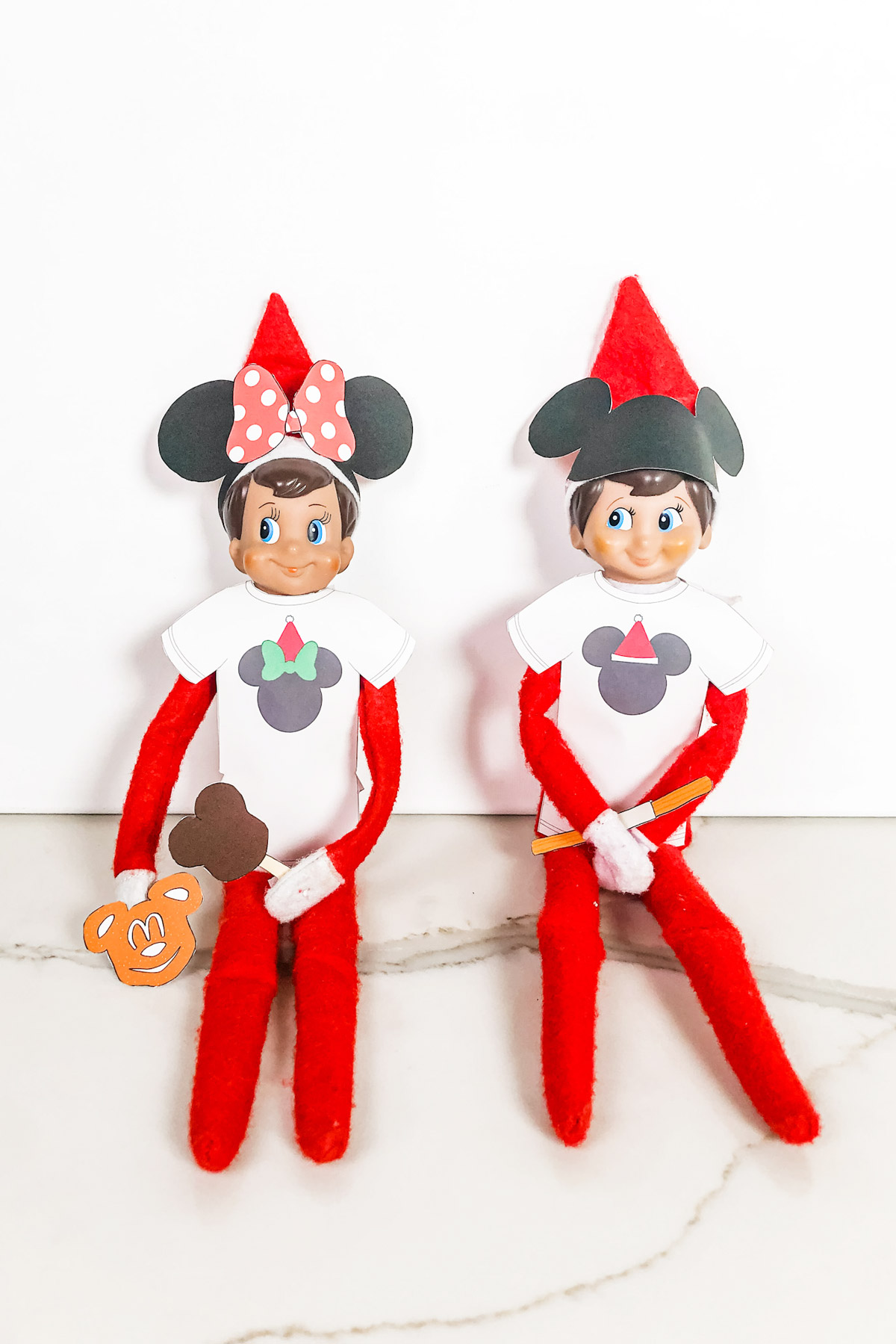 This image shows two Elf on the Shelf dolls wearing their free Elf on the Shelf Mouse Ears and Snacks printable items that you can get for free at the end of this blog post.