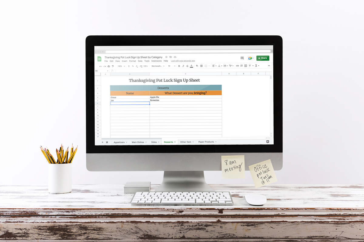 This image shows one of the two Google Sheets options for the free Thanksgiving potluck sign up sheet set you can download at the end of this post. The image shows a computer on a wooden desk with pencils next to it. It has two post its on the bottom right of the computer - one says 8 am meeting the other says office pot luck Tues @ 11 am. The screen is on the Thanksgiving Pot Luck Sign Up Sheet Google sheet dessert category page.