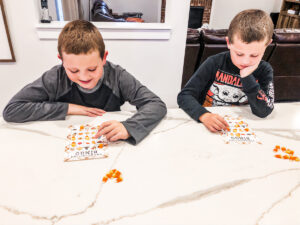 This is an image of two children playing bingo using two of the cards from the Thanksgiving bingo cards free printable set that you can get for free at the end of this blog post. They are using candy corn as their bingo markers.