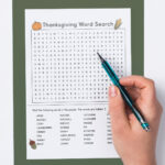This image shows a child with a pen in their hand about to complete a Thanksgiving Word search printable which you can get at the end of the blog post. This example is showing the hard version. At the top, it says Thanksgiving word search. Below that is a grid of letters. Underneath that is a list of Thanksgiving themed words to search for (such as turkey, fall, football, and more). This example also has color Thanksgiving clip art including a pumpkin, ear of corn, acorn, and slice of pumpkin pie.