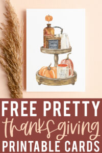 At the bottom it says free pretty Thanksgiving printable cards. Above that, the image shows one of the free Thanksgiving cards you can get from the thanksgiving cards printable set. This card has a wood tiered tray drawing with some fall items including a sign that says grateful, thankful blessed, and a mug that says hello pumpkin. It is surrounded by a stalk of wheat.
