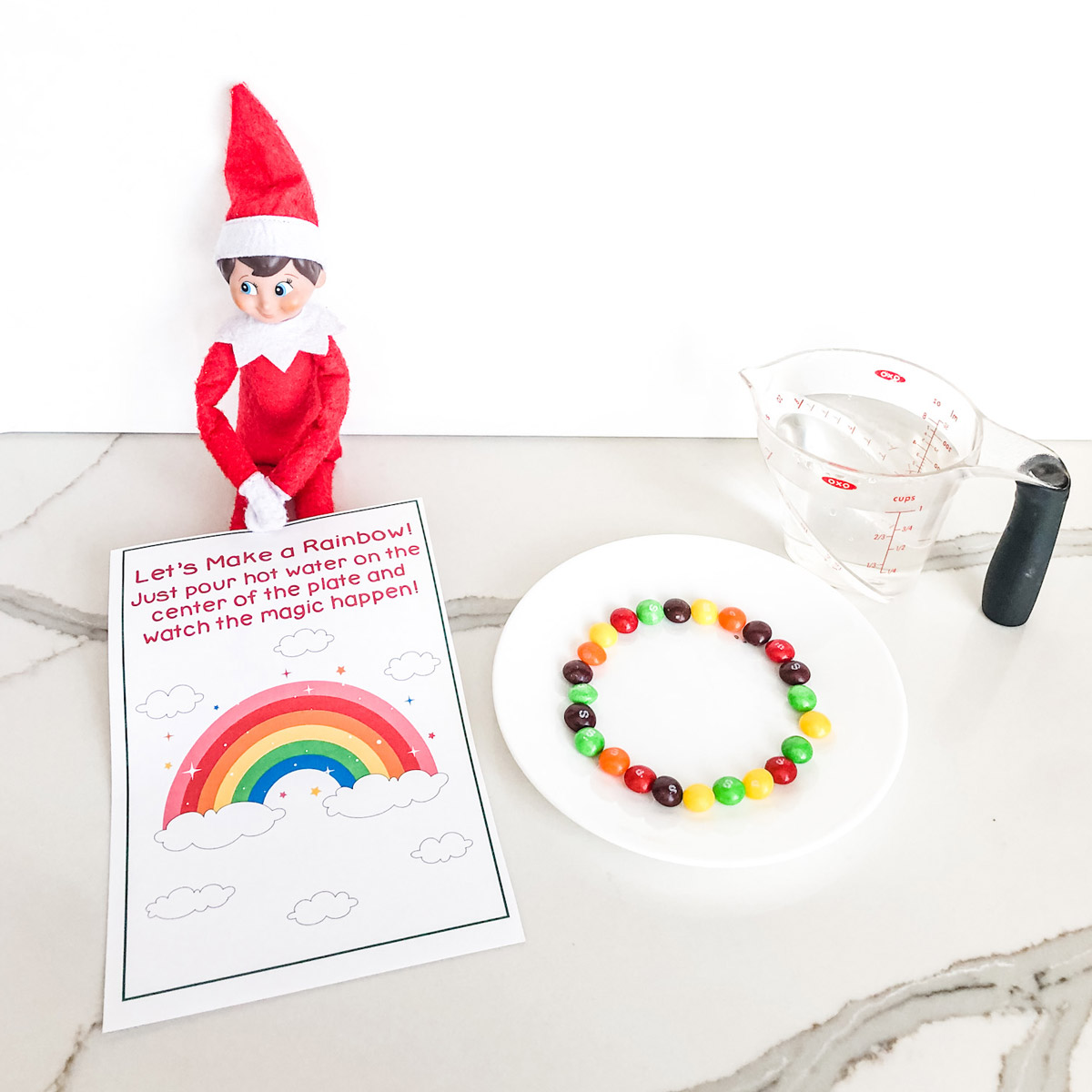 This image shows the Elf on the Shelf skittles rainbow printable that you can get for free at the end of this blog post. The Elf on the Shelf is sitting with a plate of skittles, the free printable skittles magic note and a measuring cup of water..