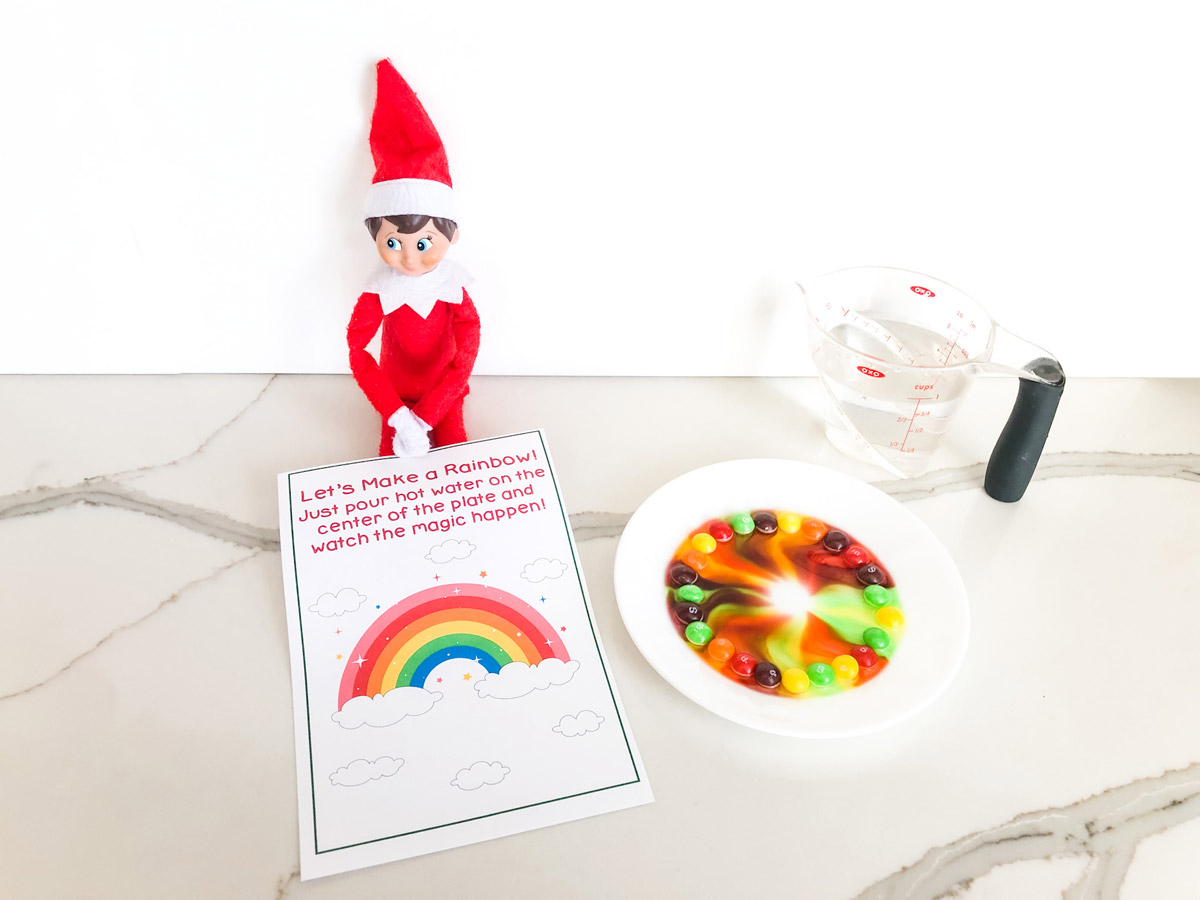 This image shows the Elf on the Shelf skittles rainbow printable that you can get for free at the end of this blog post. The Elf on the Shelf is sitting with a plate of skittles, the free printable skittles magic note and a measuring cup of water..