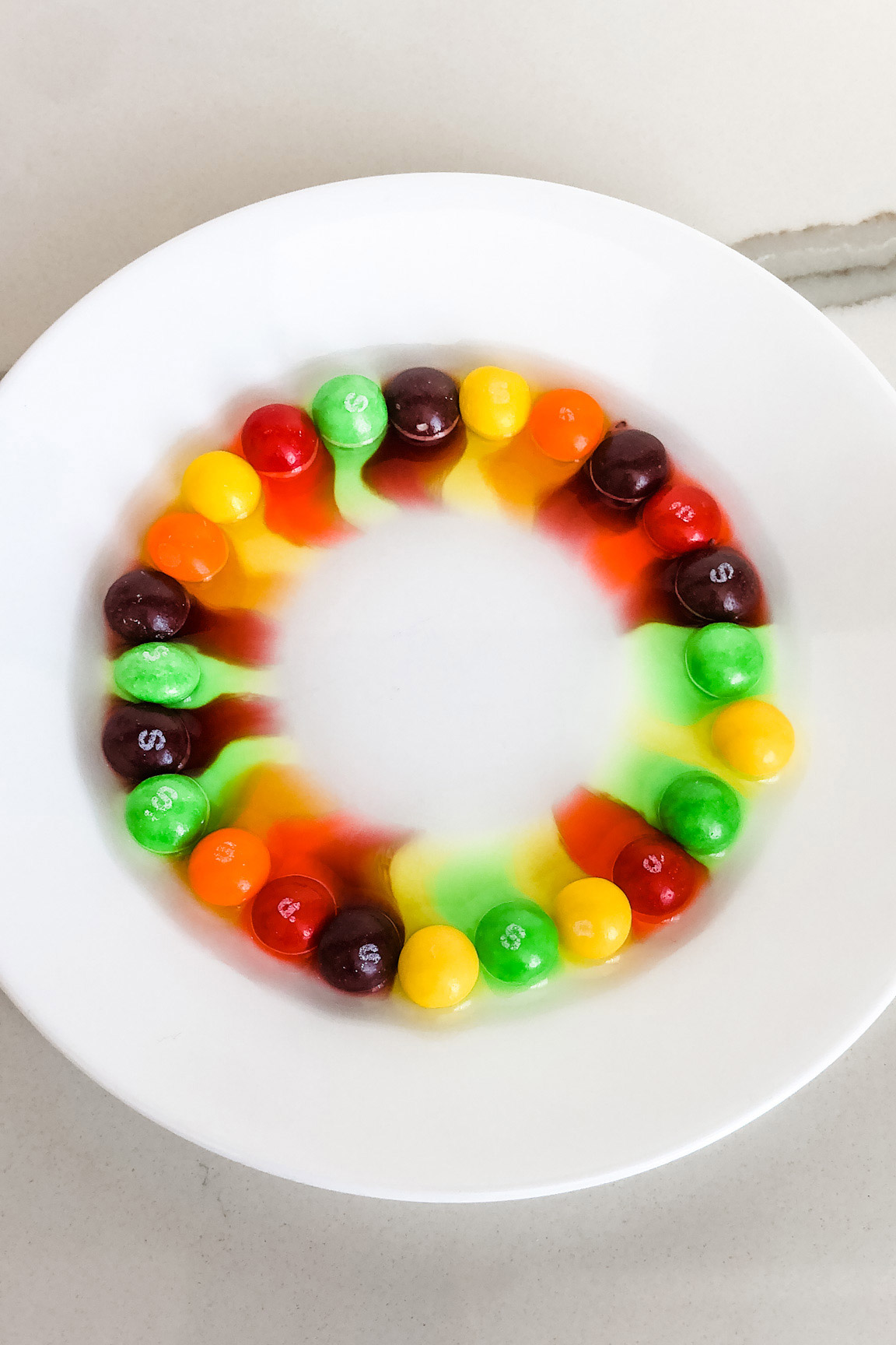 This image shows skittles on a plate that have had hot water poured on them creating a rainbow effect on the plate.