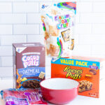 This image shows the materials you need to make the DIY I love you cerealsly bowl. It includes cereal, cereal themed oatmeal, cereal bars, a red bowl, and red silverware.