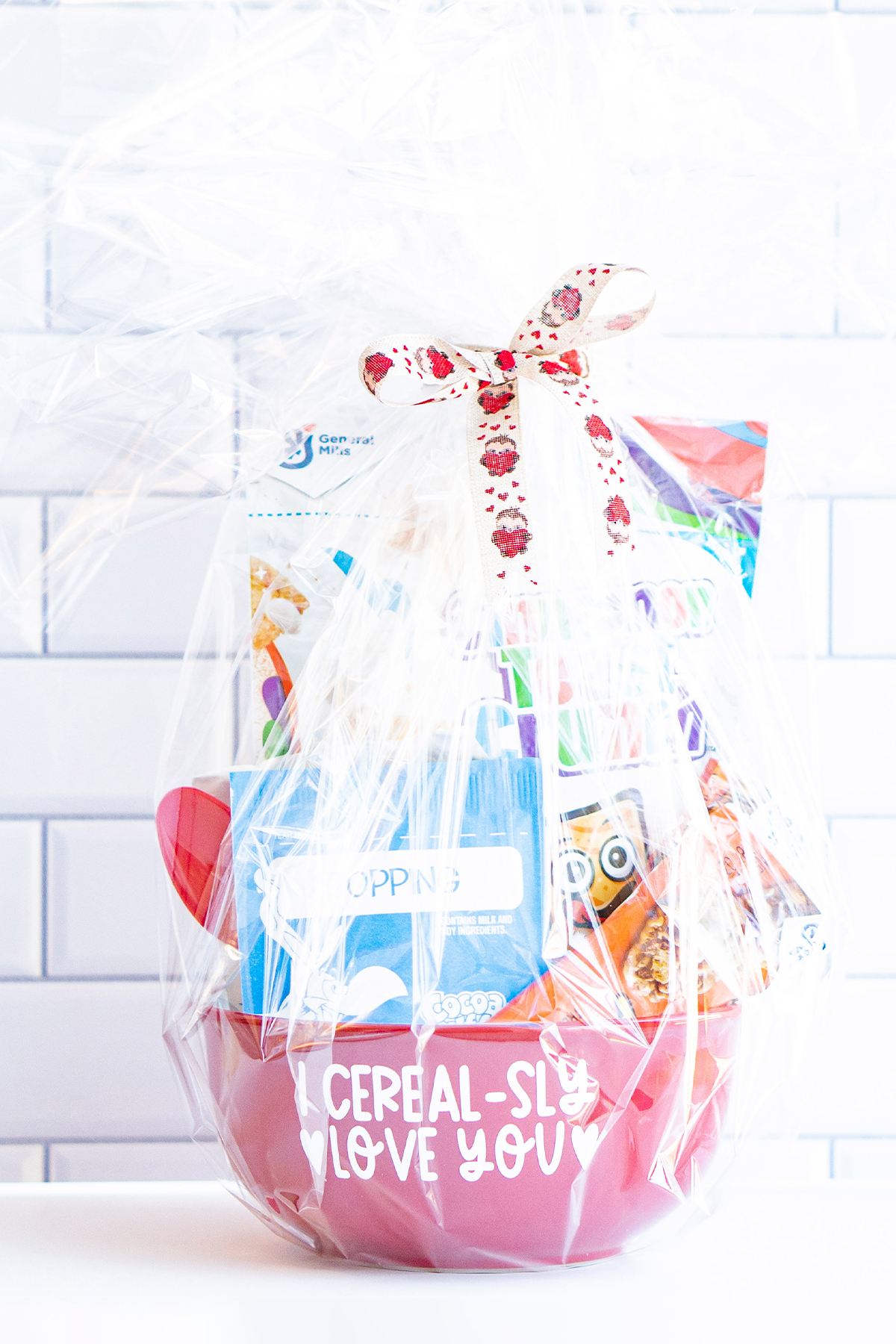 This image shows the I cerealsly love you SVG applied to a red cereal bowl. The bowl has a bag of cereal, two cereal bars, a bag of oatmeal, and a red spoon sitting inside of it. The whole thing is wrapped in clear cellophane with a ribbon at the top.