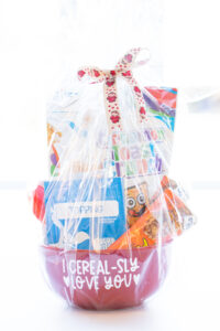 This image shows the I cerealsly love you SVG applied to a red cereal bowl. The bowl has a bag of cereal, two cereal bars, a bag of oatmeal, and a red spoon sitting inside of it. The whole thing is wrapped in clear cellophane with a ribbon at the top.