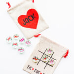 This image is showing the Valentine tic tac toe SVG you can get for free in this blog post. It is being showed on a small brown canvas bag and with small little clear gems that say x and on on them. There is also a name on the front it says Jack. The back shows a tic tac toe board with the words tic tac toe at the bottom.