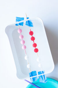 This image shows wood beads strung through wood skewers. The beads have been painted in pink, white, and red.