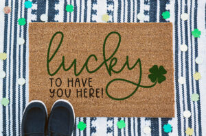 This image shows a doormat layered on top of another doormat with a pair of black shoes in the bottom left corner. Around the doormat are paper four leaf clovers and gold coins. The doormat says lucky to have you here! It's the free St. Patrick's Day svg you can get in this blog post.