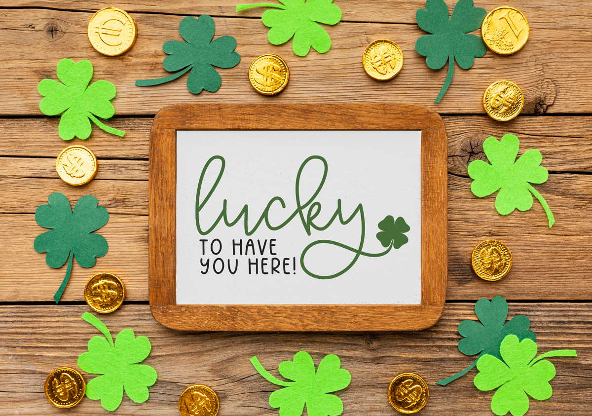 This image shows a wood sign surrounded by paper four leaf clovers and gold coins. The sign says lucky to have you here! It's the free St. Patrick's Day SVG you can get in this post.