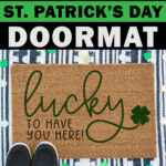At the top it says free SVG for a St. Patrick's Day doormat. Below that, the image shows a doormat layered on top of another doormat with a pair of black shoes in the bottom left corner. Around the doormat are paper four leaf clovers and gold coins. The doormat says lucky to have you here! It's the free St. Patrick's Day svg you can get in this blog post. At the bottom it says Silhouette cut file & PNG included, too.