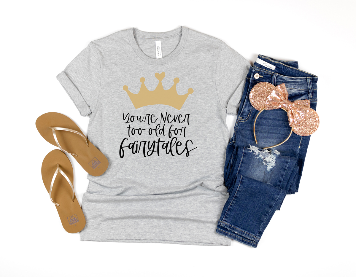 This image shows a shirt with the you're never too old for fairy tales SVG file you can get for free in this post. Next to the shirt are a pair of mouse ears on a pair of jeans and to the left is a pair of flip flops.