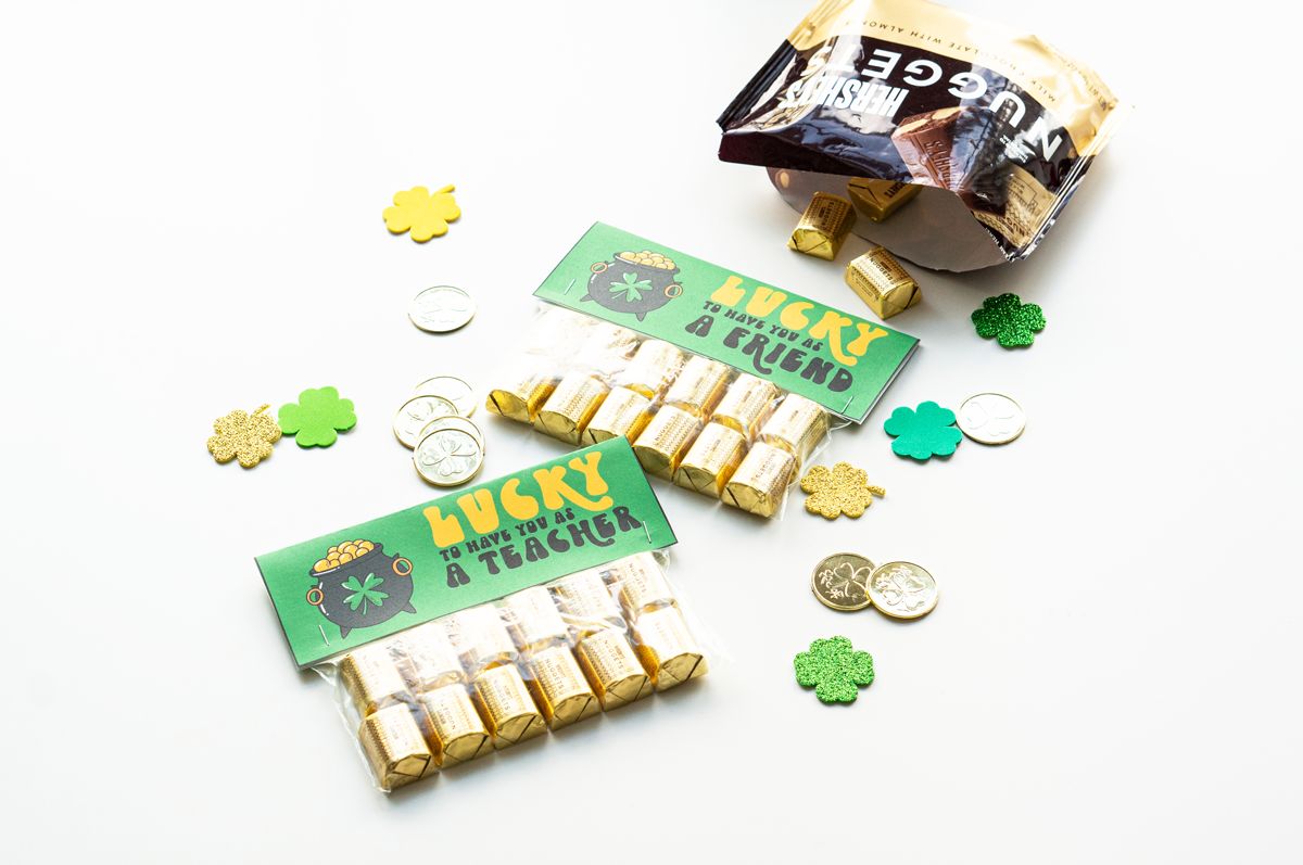 This image shows 2 completed free lucky to have you gifts. It says on it, lucky to have you as a friend on one and lucky to have you as a teacher on the other. Each topper is attached to a bag filled with Hershey's Nuggets. They are surrounded by plastic gold coins and four leaf clover cut outs. In the top right corner is a bag of Hershey Nuggets with a few on spilled onto the table.