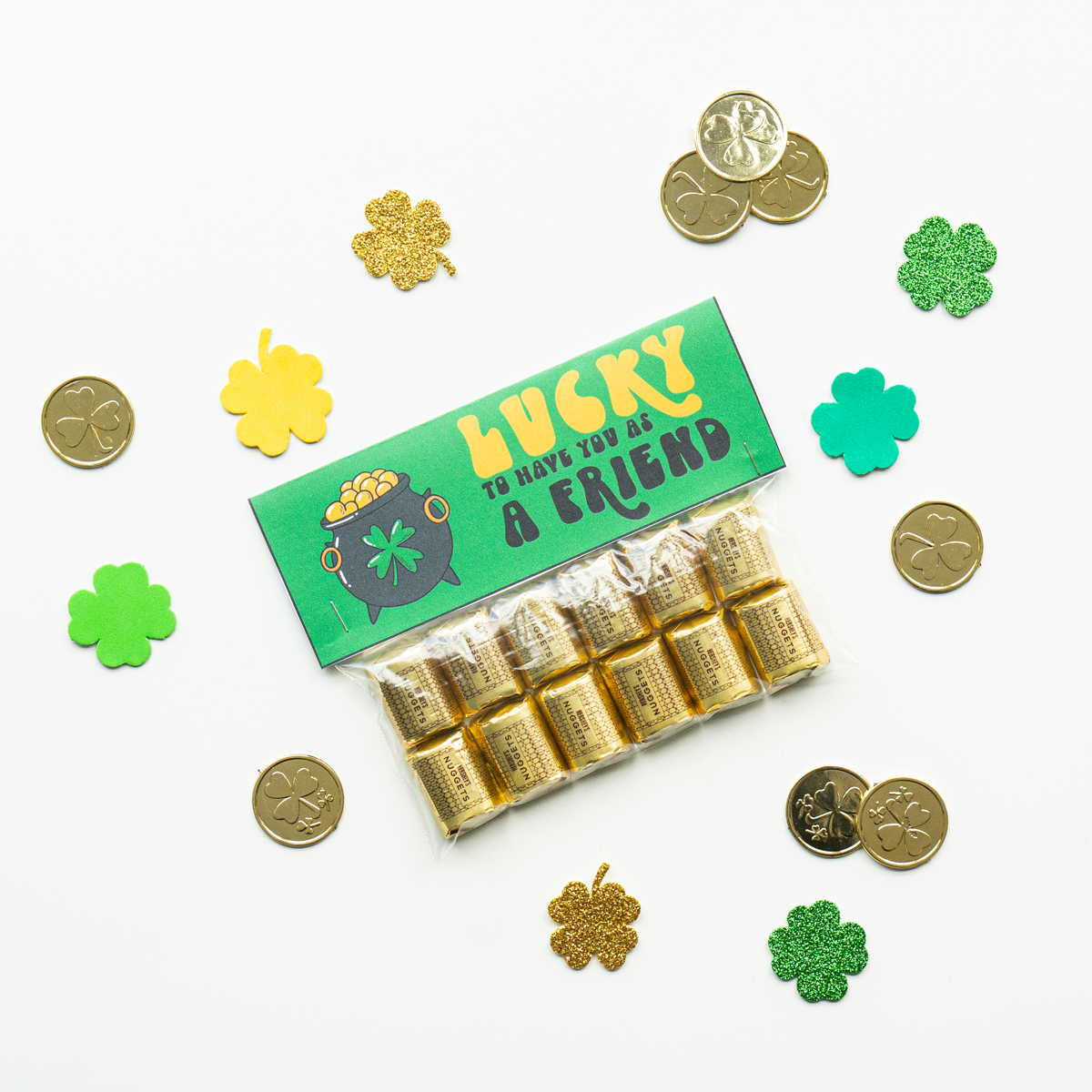 This image shows a completed free lucky to have you gift. It says on it, lucky to have you as a friend. It is attached to a bag filled with Hershey's Nuggets. It is surrounded by plastic gold coins and four leaf clover cut outs.