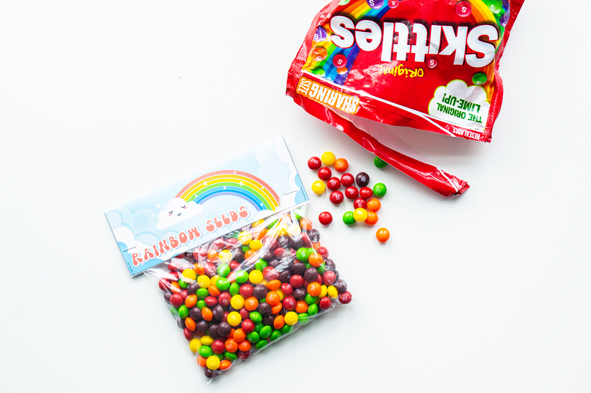 This image shows a bag of skittles with a bag topper that says rainbow seeds on it with a picture of a rainbow. In the top right corner is a bag of skittles with some skittles spilled out on the table.