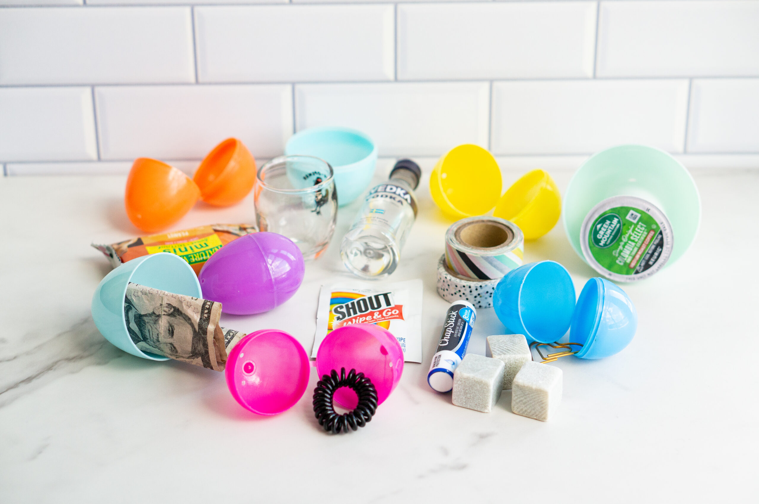 This image shows items that can fit into an Easter egg for adults, this one shows a bunch of plastic eggs and money, hair tie, whiskey rocks, chapstick, coffee pod, granola bar, shot class, washi tape, and mini liquor bottle.