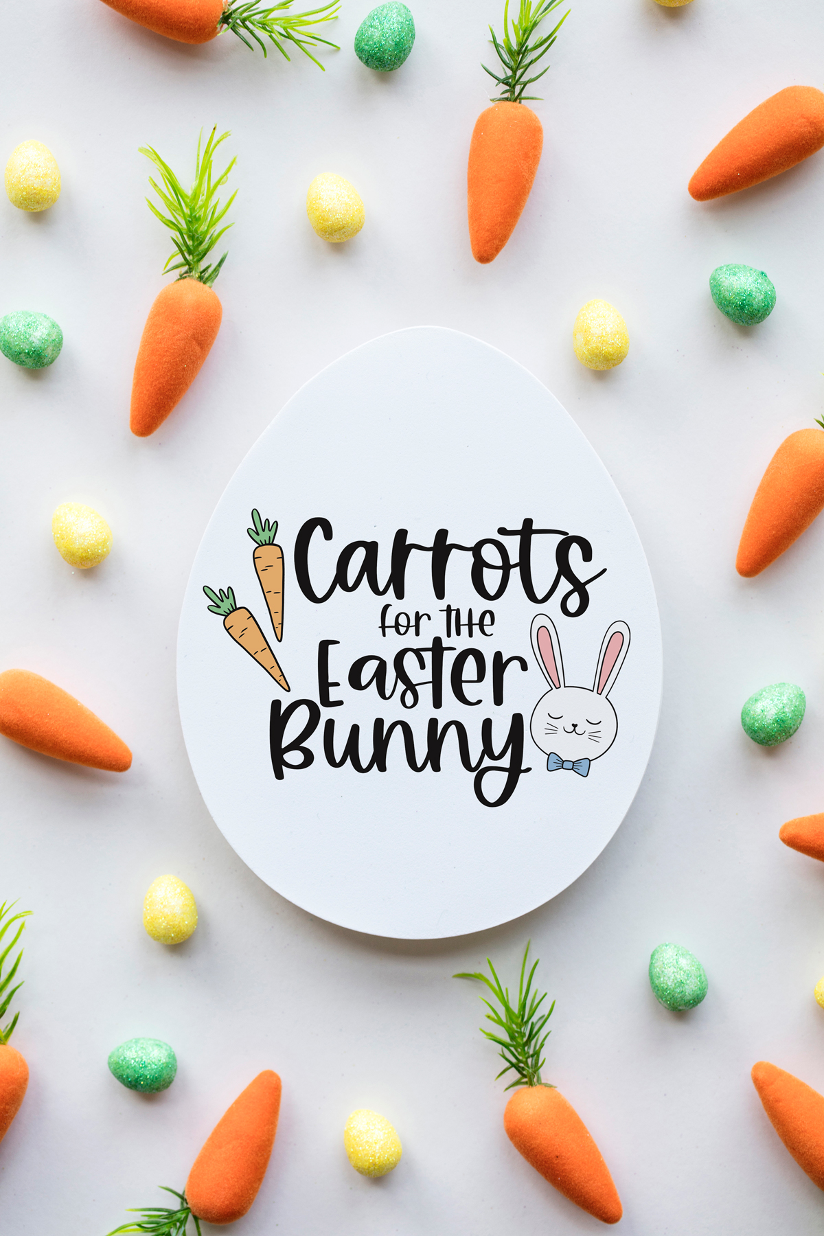 This image shows an egg shaped plate with the words Carrots for the Easter Bunny on it. It is surrounded by small color eggs and faux carrots.