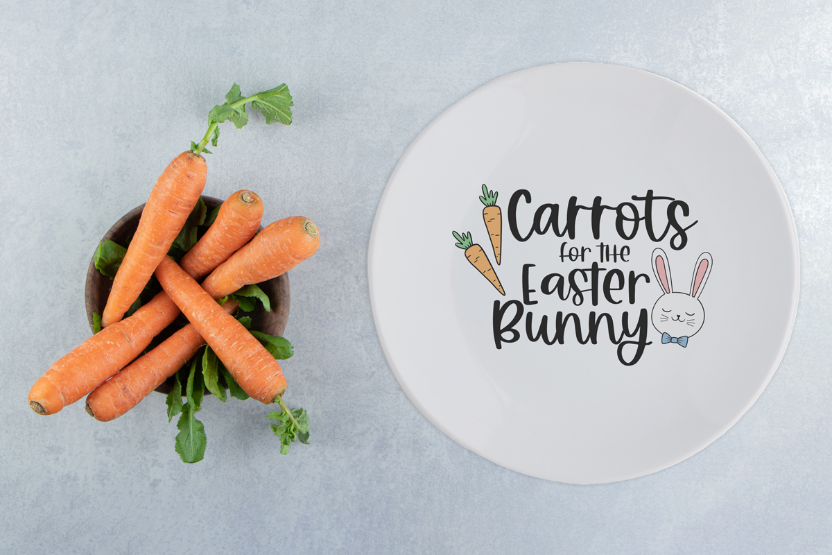 This image is of a round white plate with the words Carrots for the Easter bunny on it. There is a bin of carrots next to the plate.