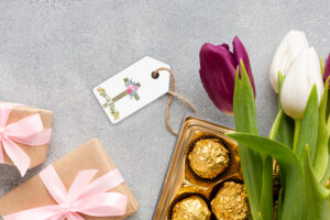 This image shows one of the free pretty Easter gift tags you can download in this post. It is next to a box of chocolates, purple and white tulips, and 2 small gift boxes with pink bows.