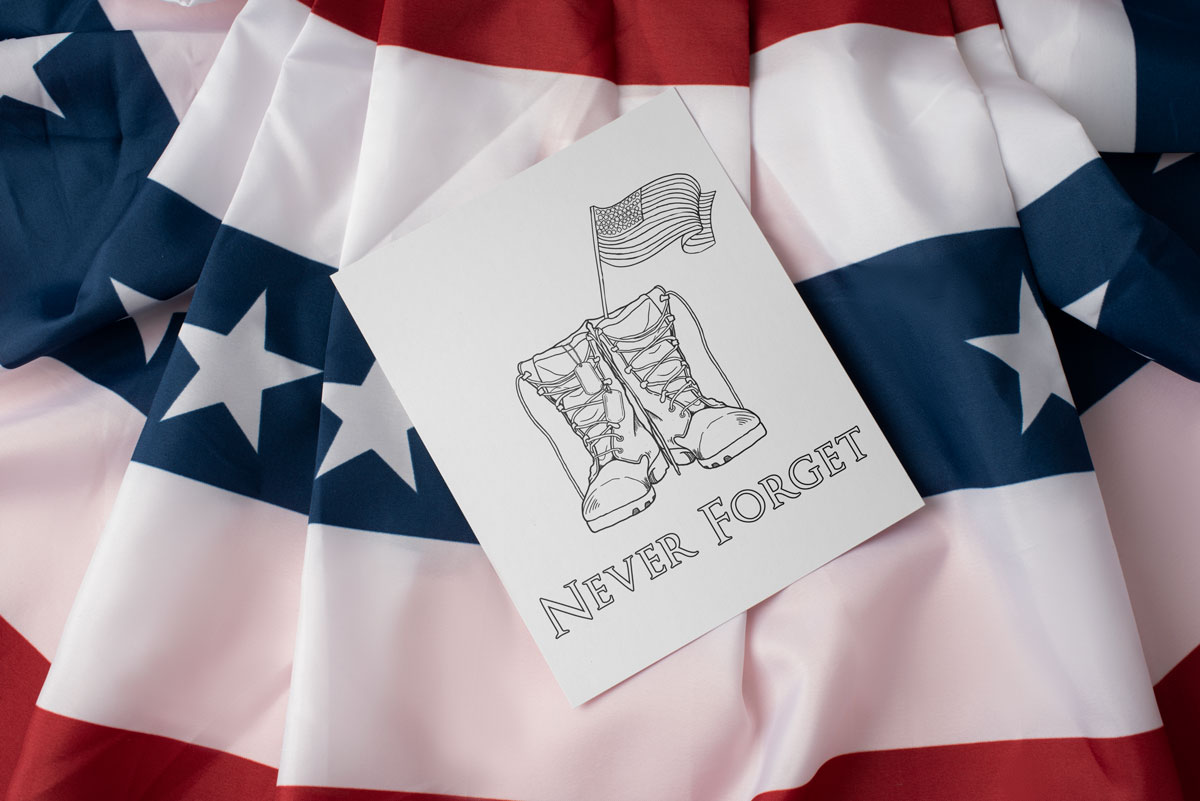 This Memorial Day coloring page shows a pair of boots that look like ones typically worn during military service. There is a pair of dog tags hanging from the left boot and an American flag coming out of the top of the right boot. Below that it says Never Forget. The coloring page is sitting on patriotic bunting fabric.