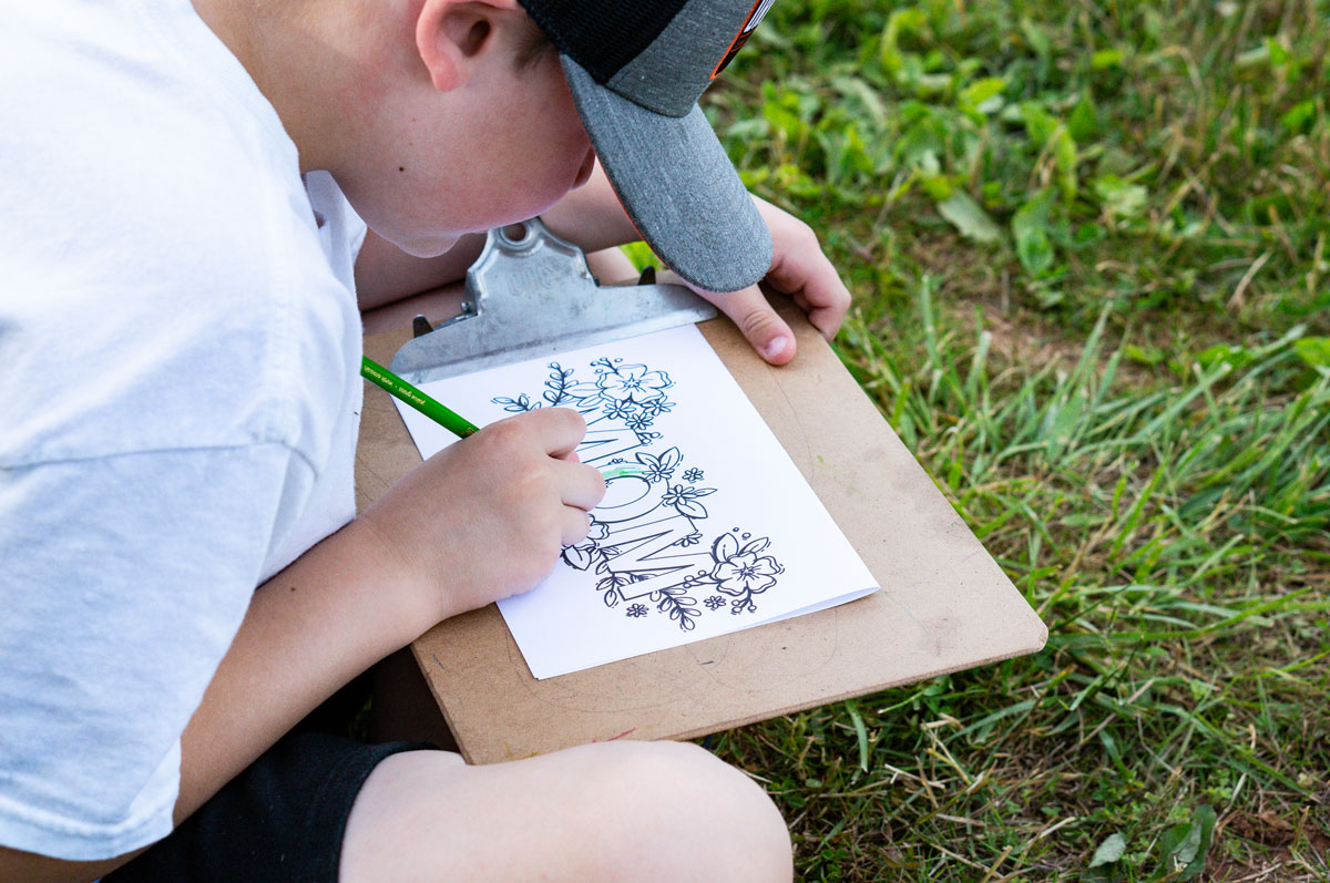 This picture shows a little boy coloring in one of the free printable Mother's Day cards to color. The card says Mom with flowers surrounding the word. The card is on a clipboard and he is coloring it in the color green.