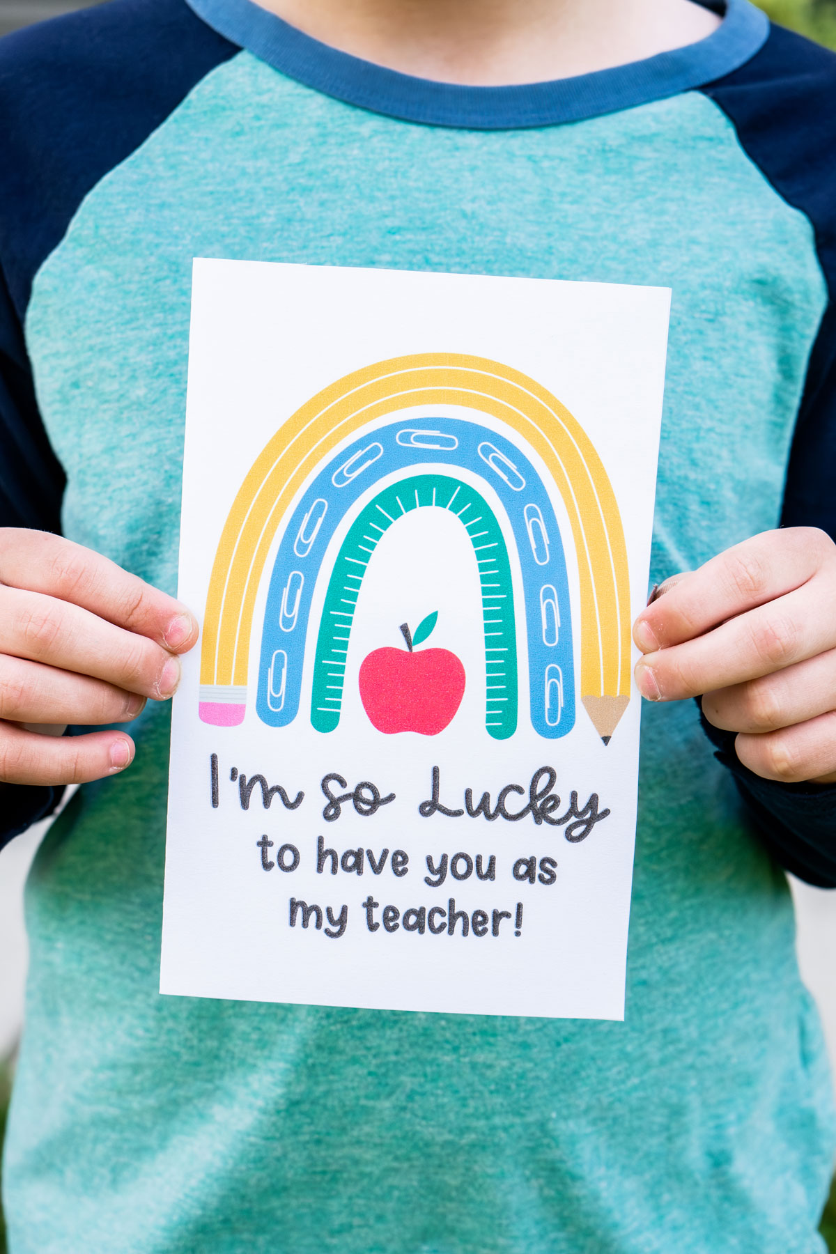 This image is of a child holding one of the free printable thank you cards for teacher appreciation or the end of the year. It says I'm so lucky to have you as my teacher! And it has a cute rainbow made out of a pencil, paper clips, and ruler.