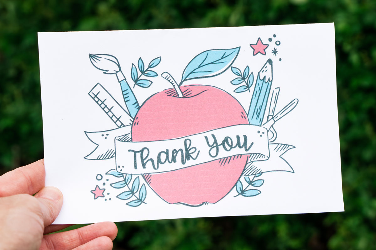 This image is of someone holding one of the free printable thank you cards for teacher appreciation or the end of the year. It says thank you and has an apple and school supplies.