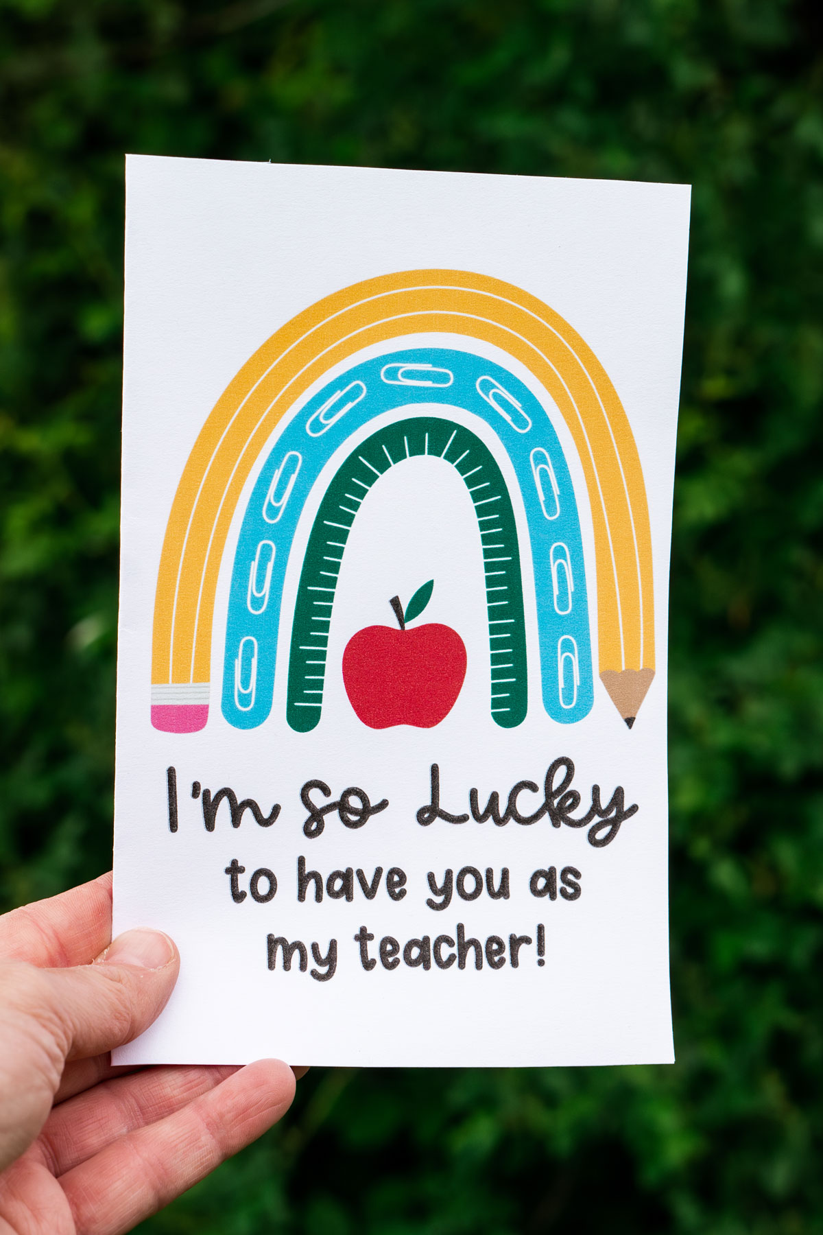 This image is of someone holding one of the free printable thank you cards for teacher appreciation or the end of the year. It says I'm so lucky to have you as my teacher! And it has a cute rainbow made out of a pencil, paper clips, and ruler.