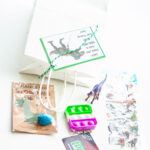 This image is showing one of the birthday party gift bag ideas which is themed for dinosaurs - dinosaur egg, dinosaur mini golf gift card, fidget, dinosaur toy, and dinosaur stickers.