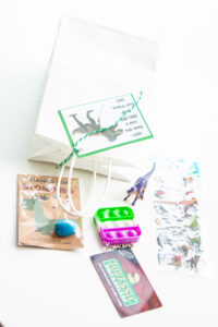 This image is showing one of the birthday party gift bag ideas which is themed for dinosaurs - dinosaur egg, dinosaur mini golf gift card, fidget, dinosaur toy, and dinosaur stickers.