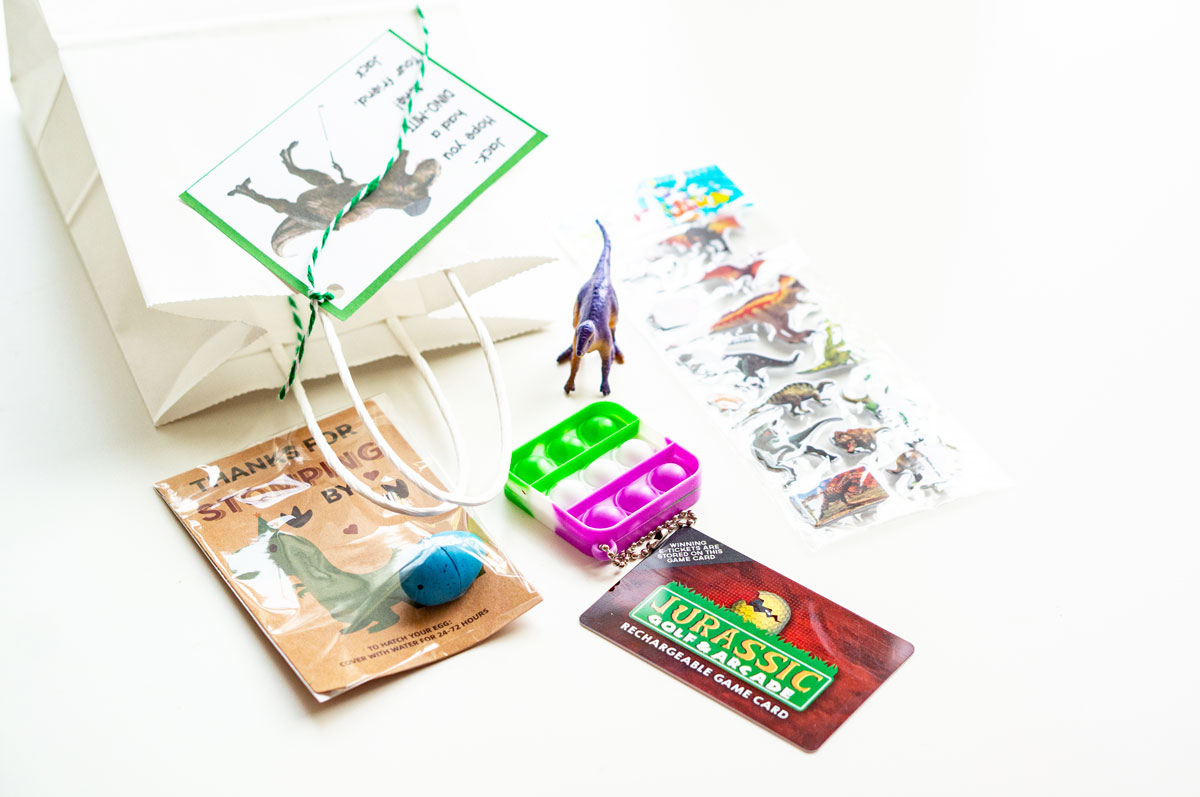 This image is showing one of the birthday party gift bag ideas which is themed for dinosaurs - dinosaur egg, dinosaur mini golf gift card, fidget, dinosaur toy, and dinosaur stickers.