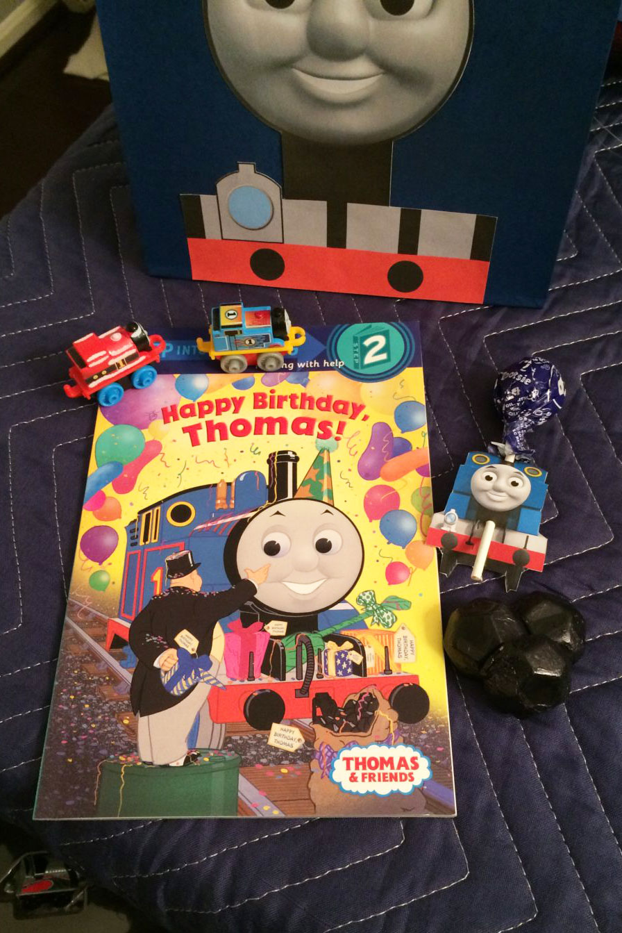 This image is showing one of the birthday party gift bag ideas which is a Thomas the Tank Engine train themed bag with a Thomas book, toy trains, coal stress ball, and a lollipop.