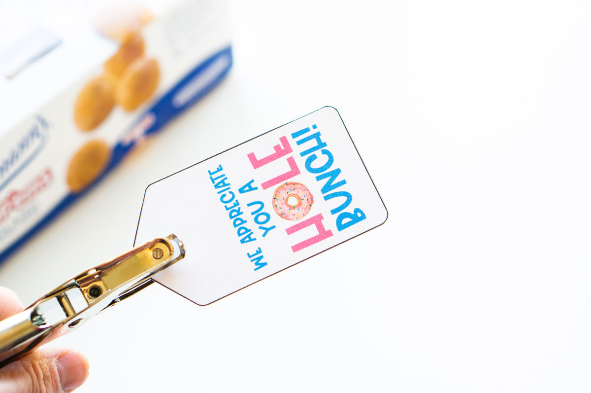 This image shows hole punching one of the free teacher appreciation tags - it says we appreciate you a hole bunch with the picture of a donut on it.