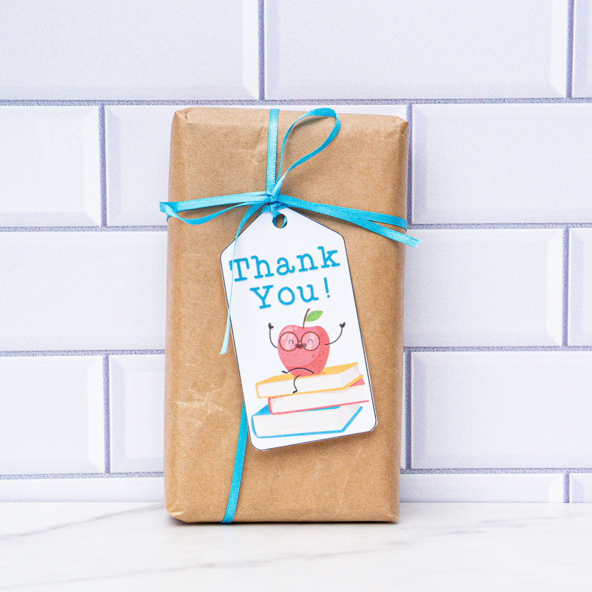 This is a box wrapped in brown kraft paper with a blue ribbon and a gift tag that says thank you with an apple sitting on a stack of books.